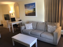 Camera d'albergo con divano grigio, una scrivania bianca, una televisione e un quadro con una spiaggia.