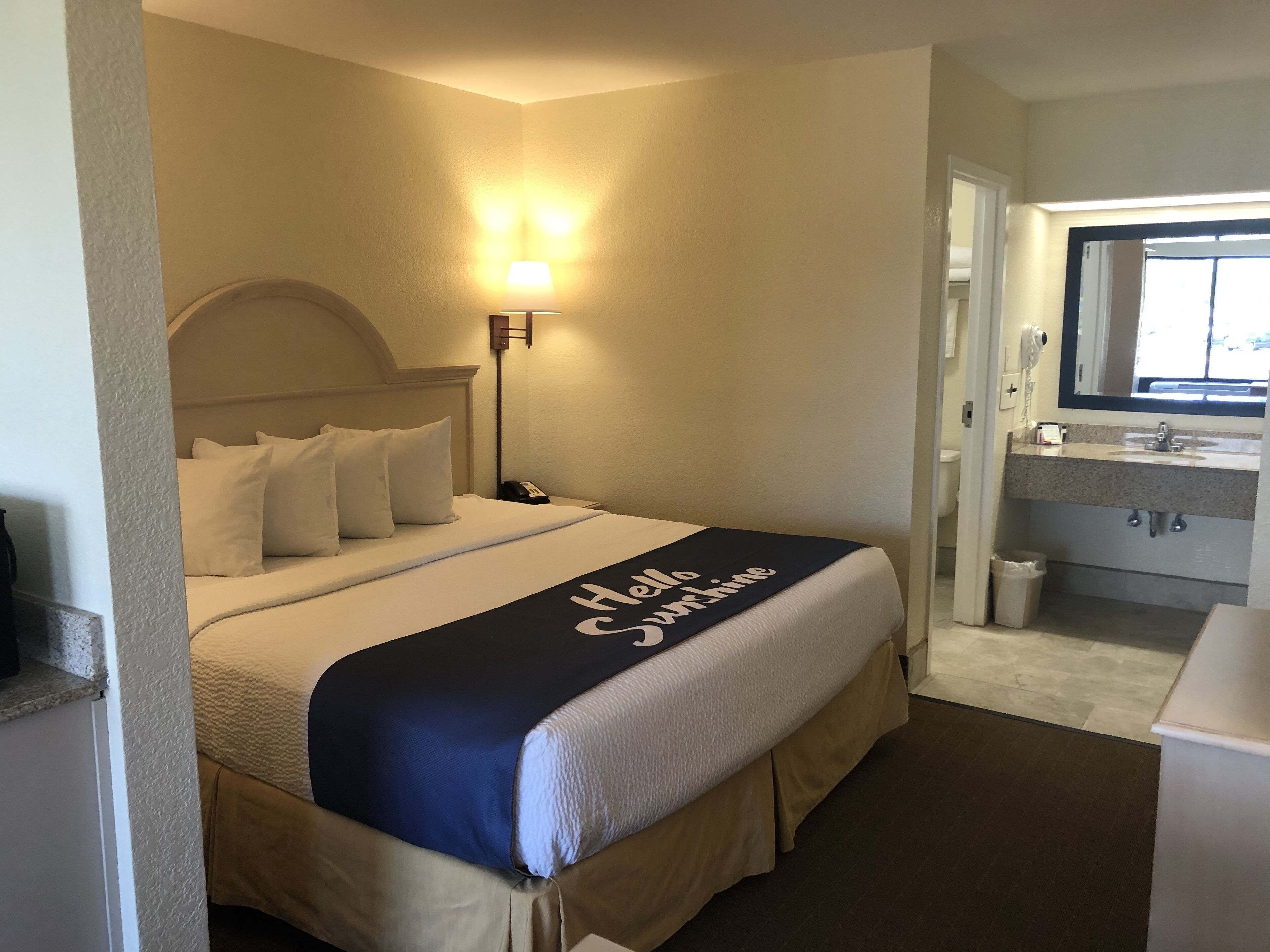 Une chambre d’hôtel avec un lit, une table de chevet, une lampe et une salle de bain équipée d’un lavabo et d’un miroir.