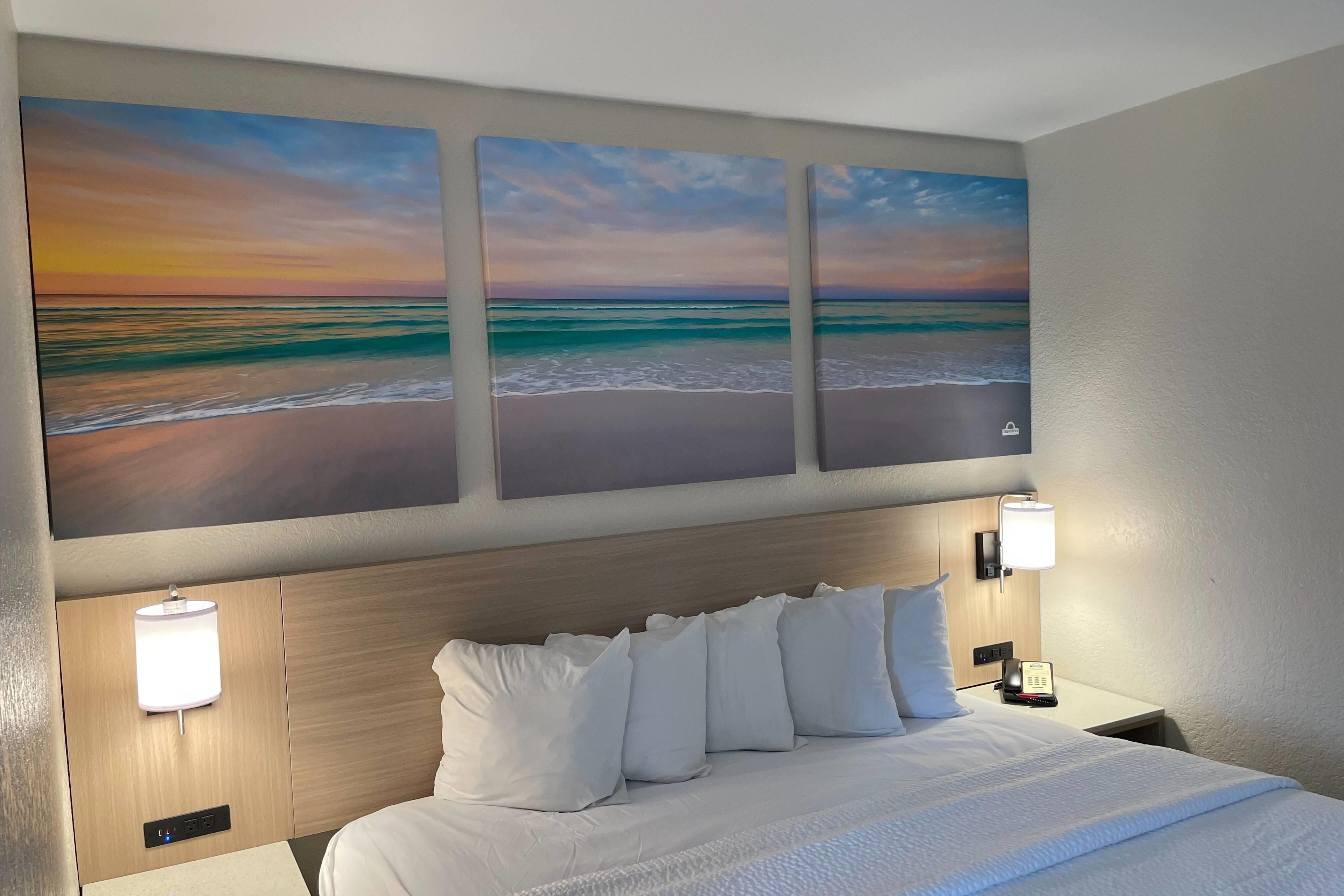 A hotel room with a bed, a headboard, wall-mounted lamps, and three paintings of a beach scene.