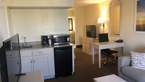 Ein kompaktes Hotelzimmer mit Küchenzeile, Schreibtisch mit Stuhl, Fernseher und einer Couch mit Zierkissen.