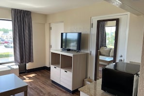 A hotel room with a television on a wooden stand, a small table, and a view of a parking lot through the window. - Days Inn & Suites by Wyndham Navarre - near Beaches/Hurlburt (Navarre)