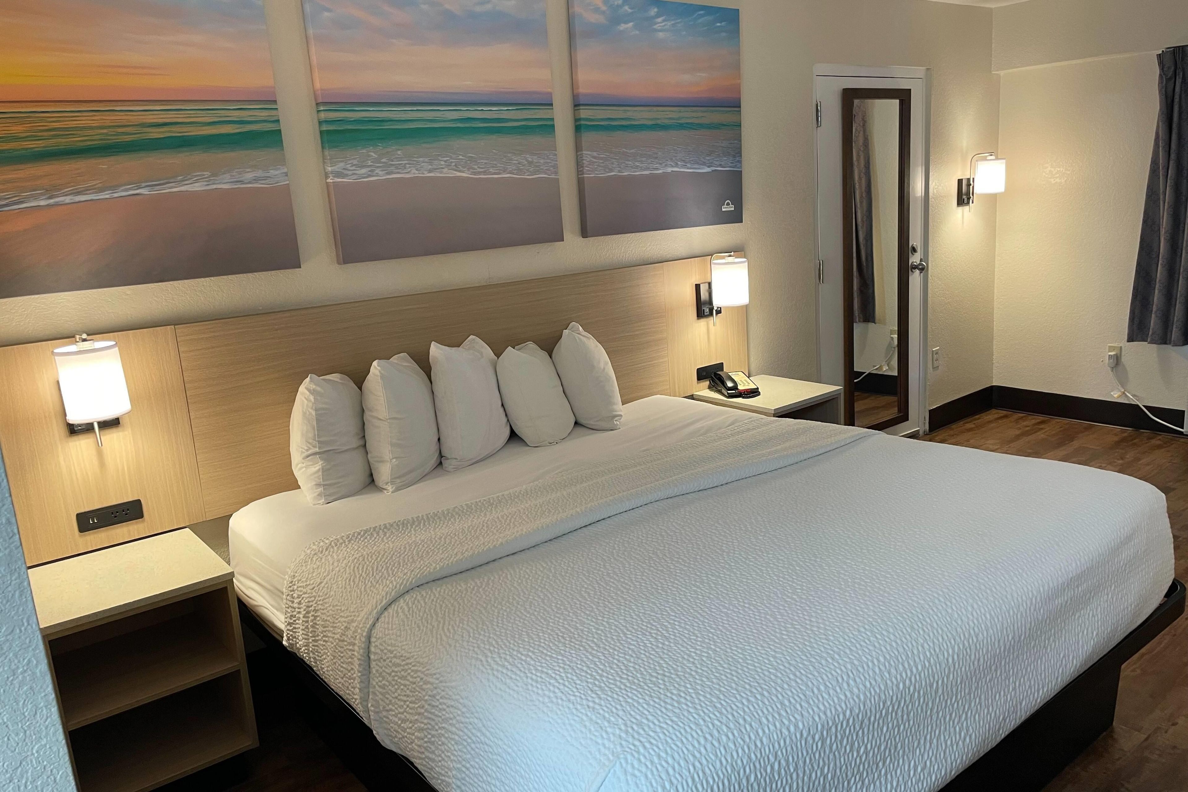 A hotel room with a large bed, a nightstand, a lamp, a phone, and a painting of a beach scene.