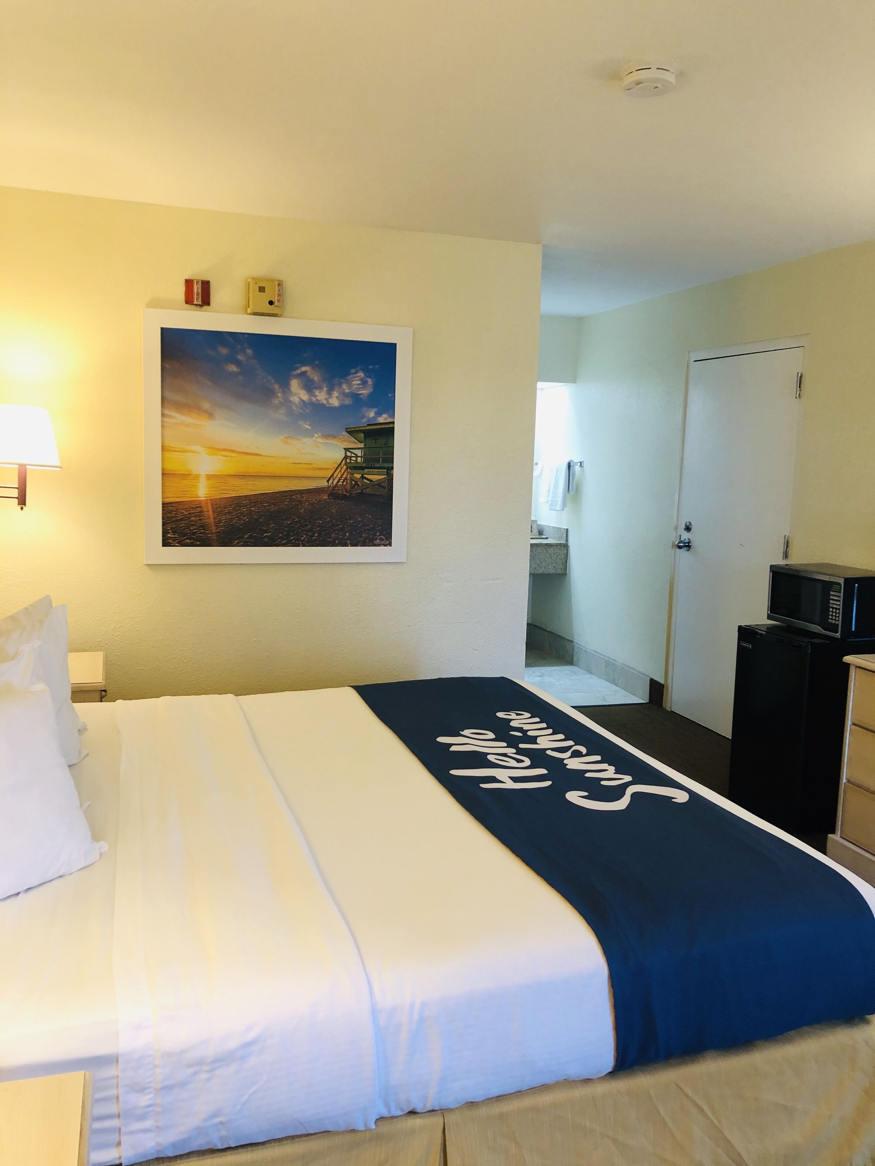 Ein Hotelzimmer mit Bett, einem Gemälde von einem Sonnenuntergang über einem Strand, einer Mikrowelle und einer Tür zu einem Badezimmer.