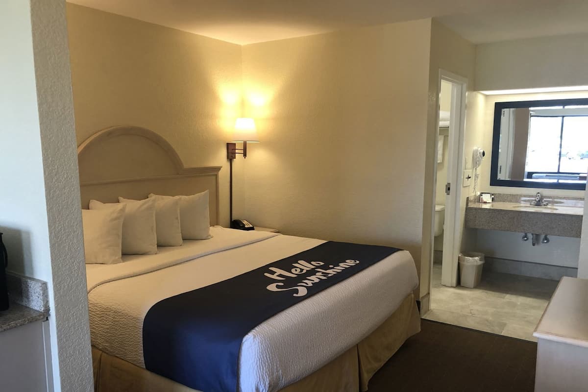 Ein Hotelzimmer mit Bett, Nachttischlampe, Badezimmer mit Waschbecken und Spiegel sowie einem Flur.
