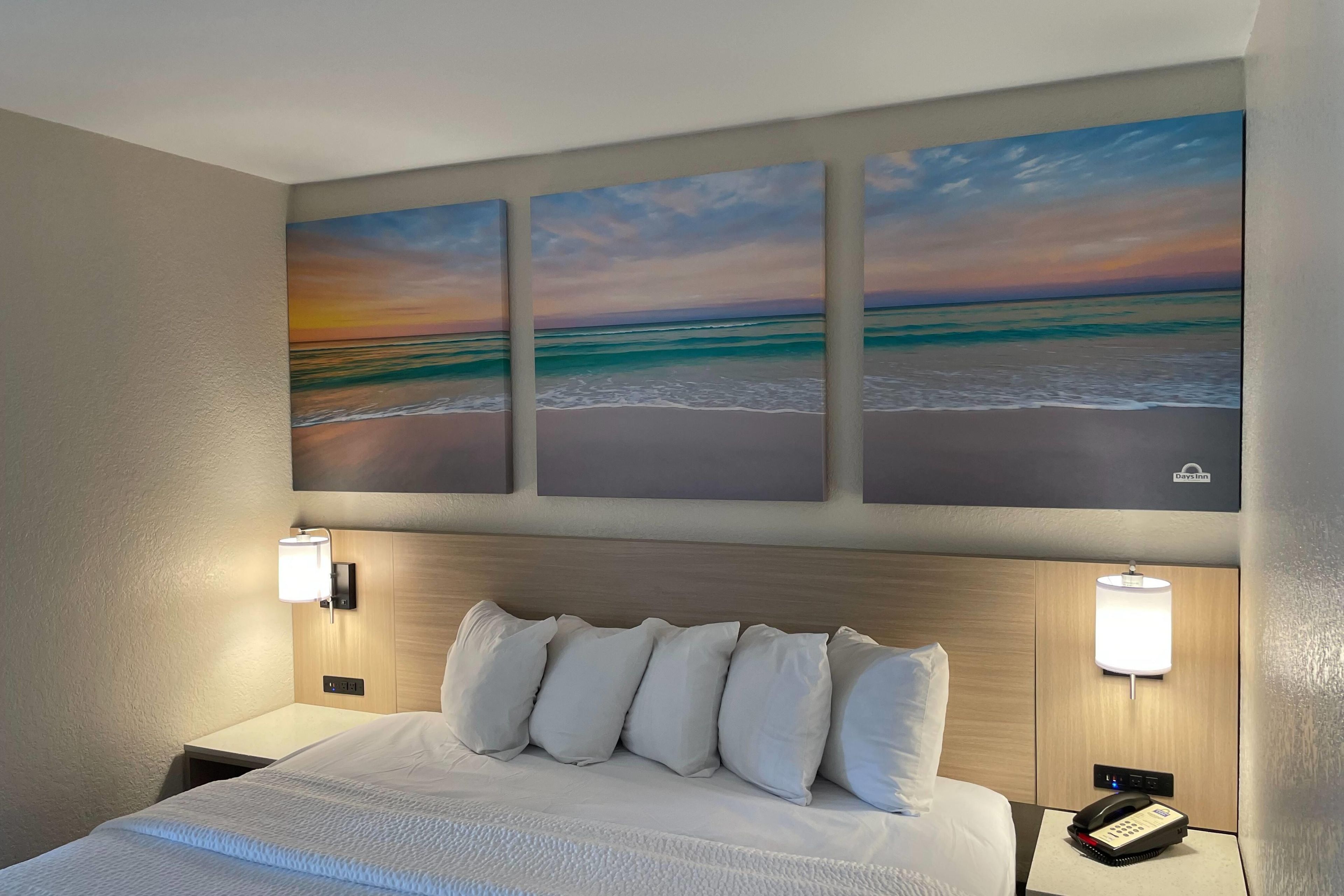 A hotel room with a bed, two bedside lamps, a nightstand, and three wall-mounted paintings of a beach scene.