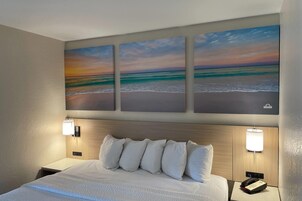 Habitación de hotel con cama, dos lámparas de noche, una mesita de noche y tres cuadros de playa colgados en la pared.