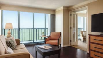 A hotel room with a sofa, armchair, coffee table, and a TV. There is a balcony with a view.