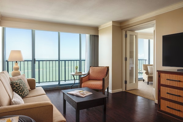 Une chambre d’hôtel équipée d’un canapé, d’un fauteuil, d’une table basse et d’un téléviseur. Elle dispose également d’un balcon avec vue.