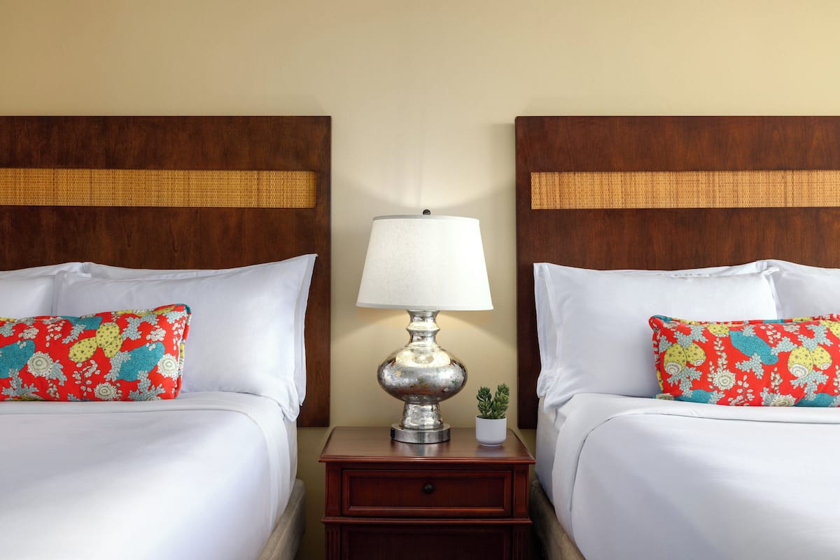 Een hotelkamer met twee bedden, elk met een witte spreidek en een kleurrijk kussen. Daartussen bevindt zich een nachtkastje met een lamp en een kleine potplant.