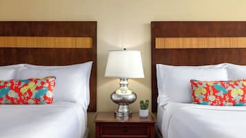A hotel room with two beds, each with a white bedspread and a colorful pillow. There is a nightstand with a lamp and a small potted plant between the beds.