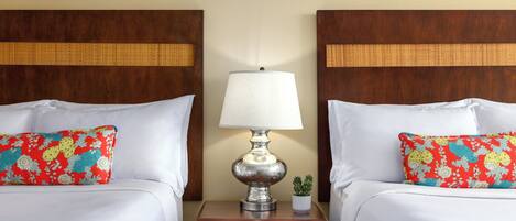 A hotel room with two beds, each with a white bedspread and a colorful pillow. There is a nightstand with a lamp and a small potted plant between the beds.