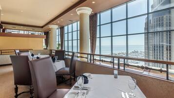 A dining area with a table set for two, large windows offering a view of the city and the sea, and a high ceiling with recessed lighting.