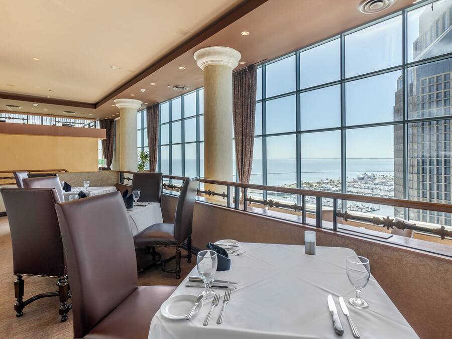 A dining area with a table set for two, large windows offering a view of the city and the sea, and a high ceiling with recessed lighting.