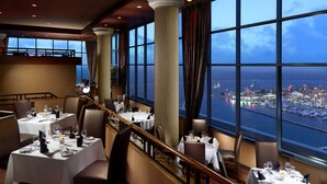 Un restaurante con mesas preparadas para comer, ventanas amplias con vistas al mar y al paisaje urbano, y un balcón con vistas al agua.