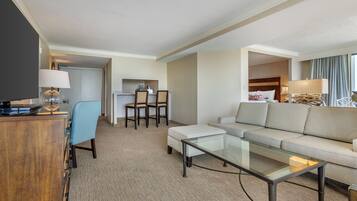 A hotel room with a sofa, a glass coffee table, a TV, a desk with a lamp, and a dining area with chairs and a table.