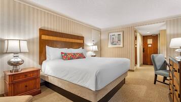 A hotel room with a large bed, a wooden headboard, a nightstand with a lamp, a chair, and a dresser.