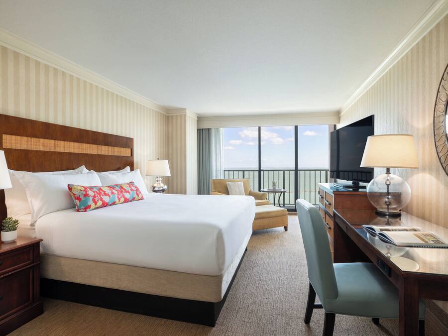 A hotel room with a large bed, a desk, a chair, a TV, and a balcony with a view.