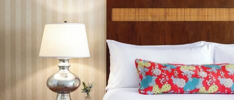 Una cama bien hecha con sábanas blancas y una almohada floral de colores, una lámpara de mesa clásica y una pequeña maceta con una planta sobre una mesita de noche de madera.