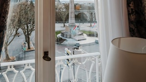 Uitzicht vanuit een raam met gordijnen, met een monument, vlaggen en een verre toren.