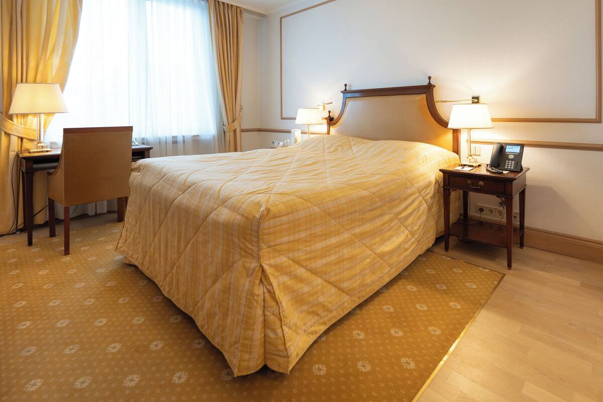 Ein Hotelzimmer mit Bett, Schreibtisch, Stuhl, Nachttischen, Lampen und Telefon.