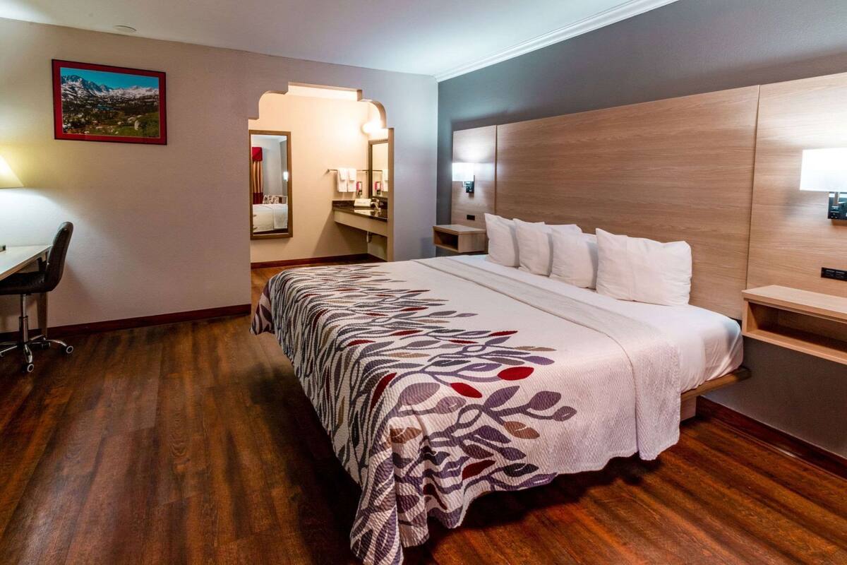 Habitación de hotel con una cama grande, un escritorio, una silla, piso de madera, una mesita de noche y una lámpara de pared.