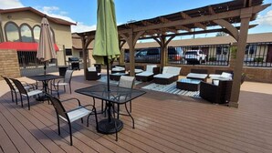 En udendørs patio med borde og stole, et pergola med en grøn parasol, og udsigt til en bygning med et skilt, der står 'Quality Inn'.