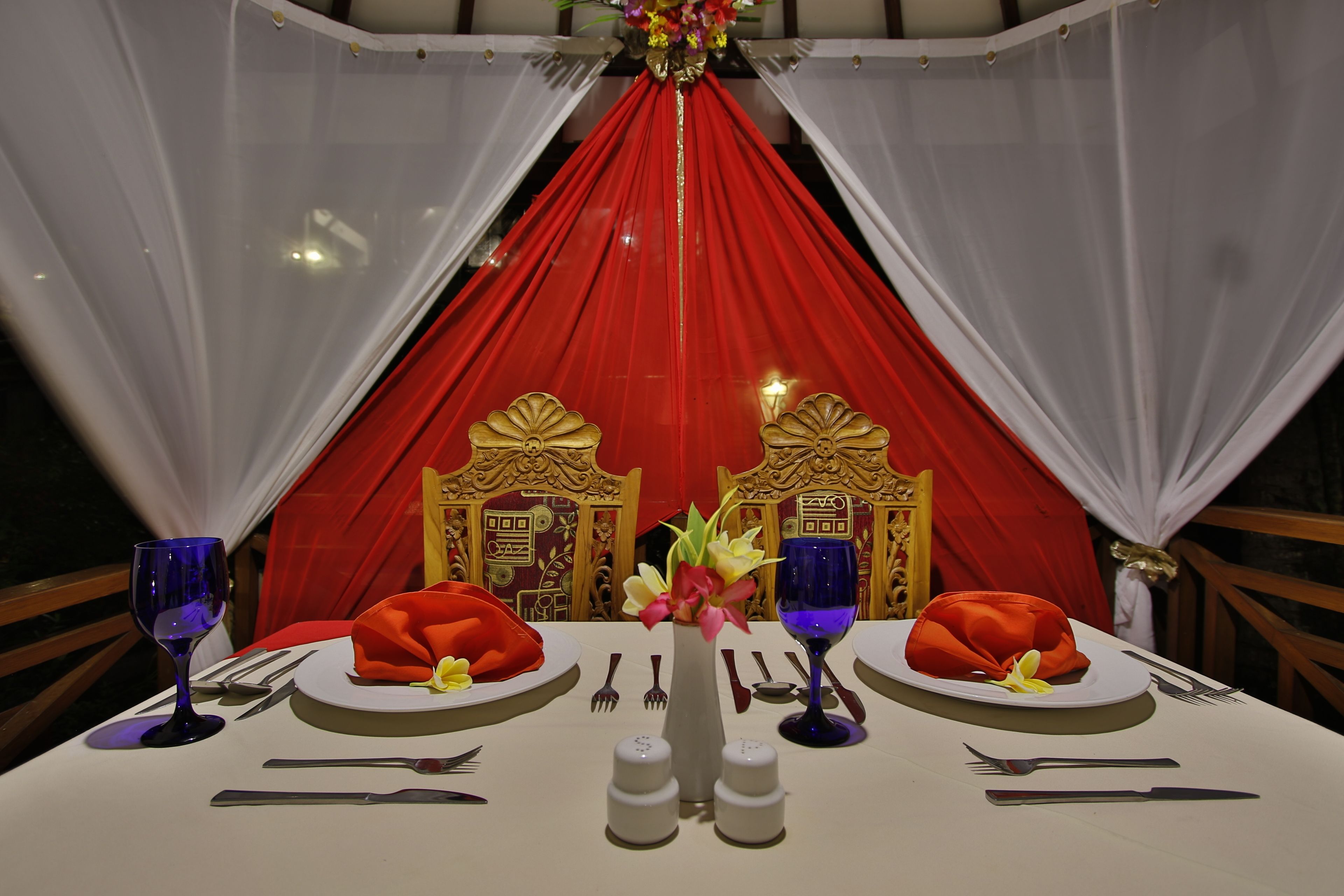 A table set for two with red and white drapery, gold accents, and a vase of flowers.