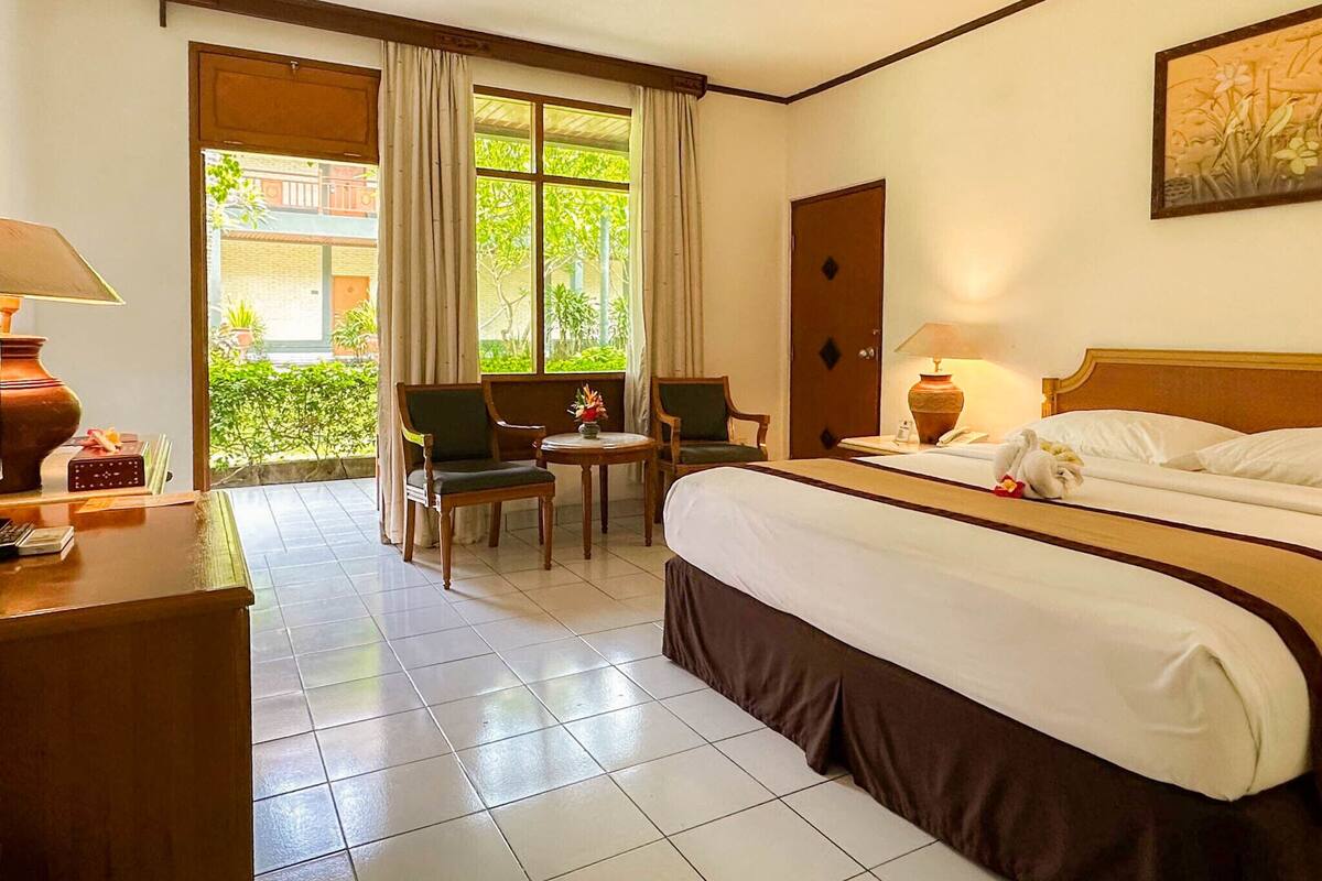 Ein Hotelzimmer mit einem Bett, zwei Sesseln, einem Schreibtisch mit Lampe, einem Fenster mit Vorhängen und einer Tür.