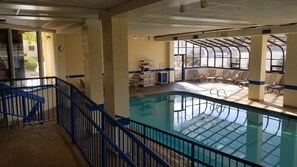An indoor swimming pool with a blue railing, a sunlit area with chairs, and a view of the outside.