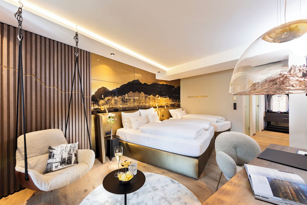 Quarto de hotel moderno com uma cama grande, uma cadeira suspensa, uma mesinha redonda com um copo e um mural com vista para a cidade na parede.