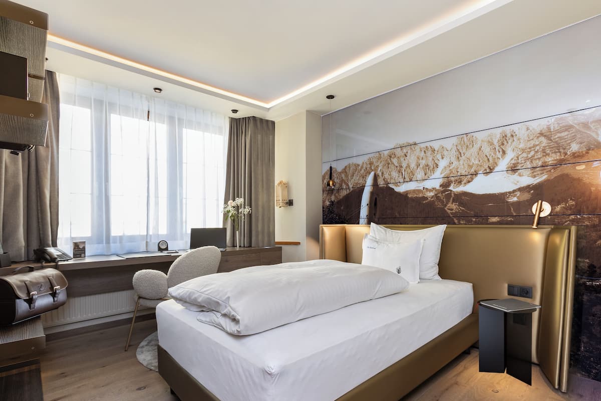 Quarto de hotel moderno com uma cama grande, uma escrivaninha com um laptop, uma cadeira e vista para uma paisagem montanhosa pela janela.