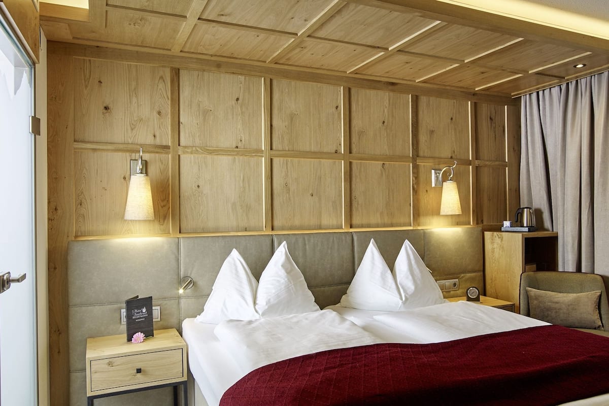 Quarto de hotel com cabeceira de madeira, cama com lençóis brancos, manta vermelha, mesa de cabeceira com abajur, criado-mudo, cadeira e janela com cortinas.