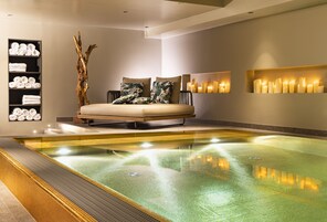 Un spa moderno con una bañera de hidromasaje grande y cerrada con vidrio, un sofá con cojines, un estante con toallas y velas fijadas a la pared.