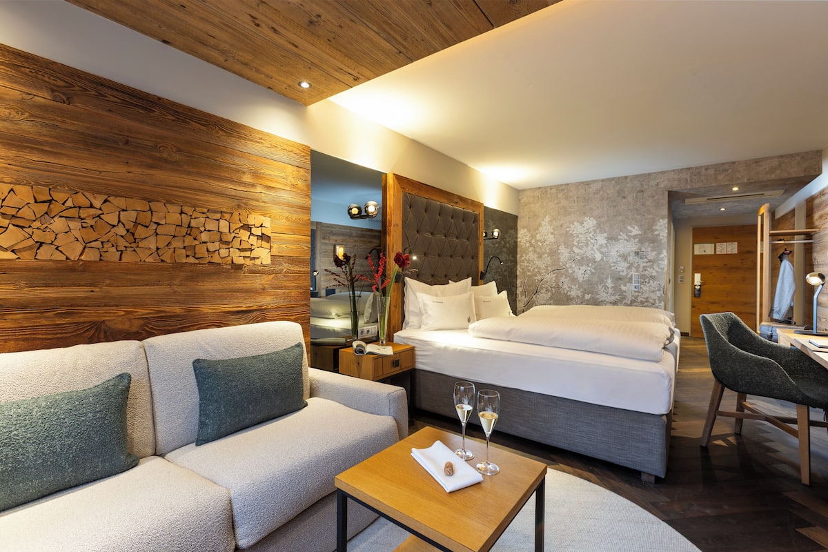 Quarto de hotel moderno com parede de destaque em madeira, uma cama grande, um sofá, uma pequena mesa com copos e uma escrivaninha com uma cadeira.