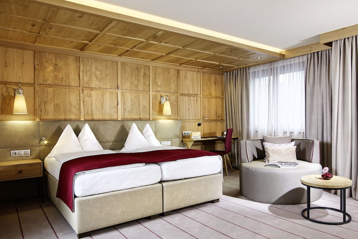 Quarto de hotel com parede de painel de madeira, cama com roupa branca, escrivaninha com cadeira e mesa lateral redonda com um vaso.