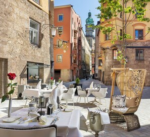 An outdoor dining area with tables set for two, surrounded by historic buildings with ornate facades and a prominent tower in the background. - Hotel Das Innsbruck (Innsbruck)