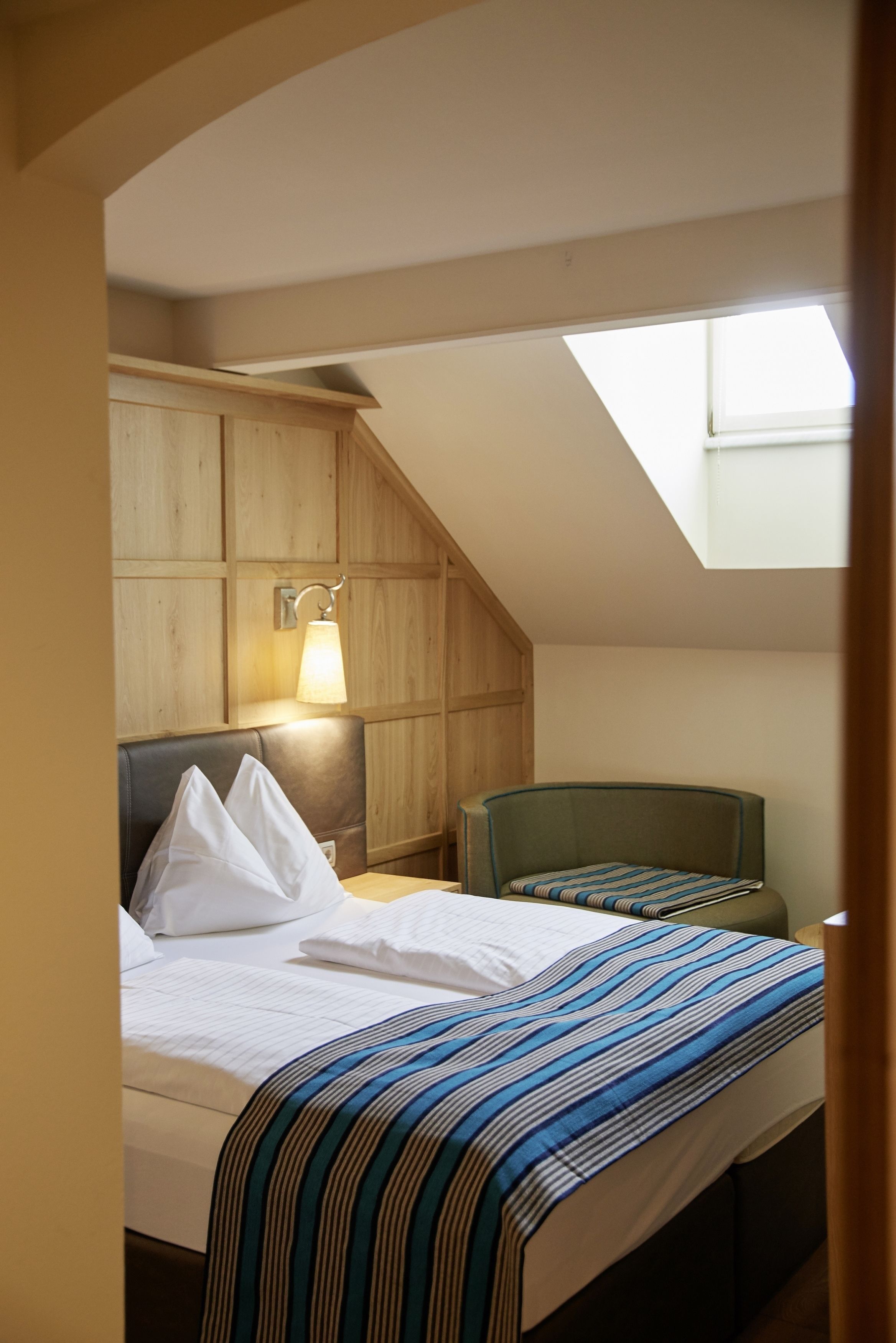 Un dormitorio con cama, mesita de noche, lámpara, silla y tragaluz.
