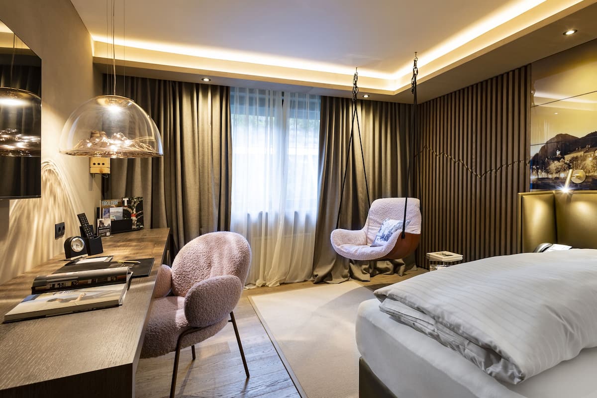 Quarto de hotel moderno com uma cama grande, uma cadeira suspensa, uma escrivaninha com abajur e vista para a paisagem urbana pela janela.