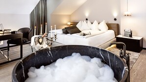 Ein modernes Hotelzimmer mit einer großen, schwarzen Badewanne voller Blasen, einem ordentlich bezogenen Bett mit weißen Leinentüchern, einem Schreibtisch mit einem Laptop und einem großen Fenster mit Vorhängen.