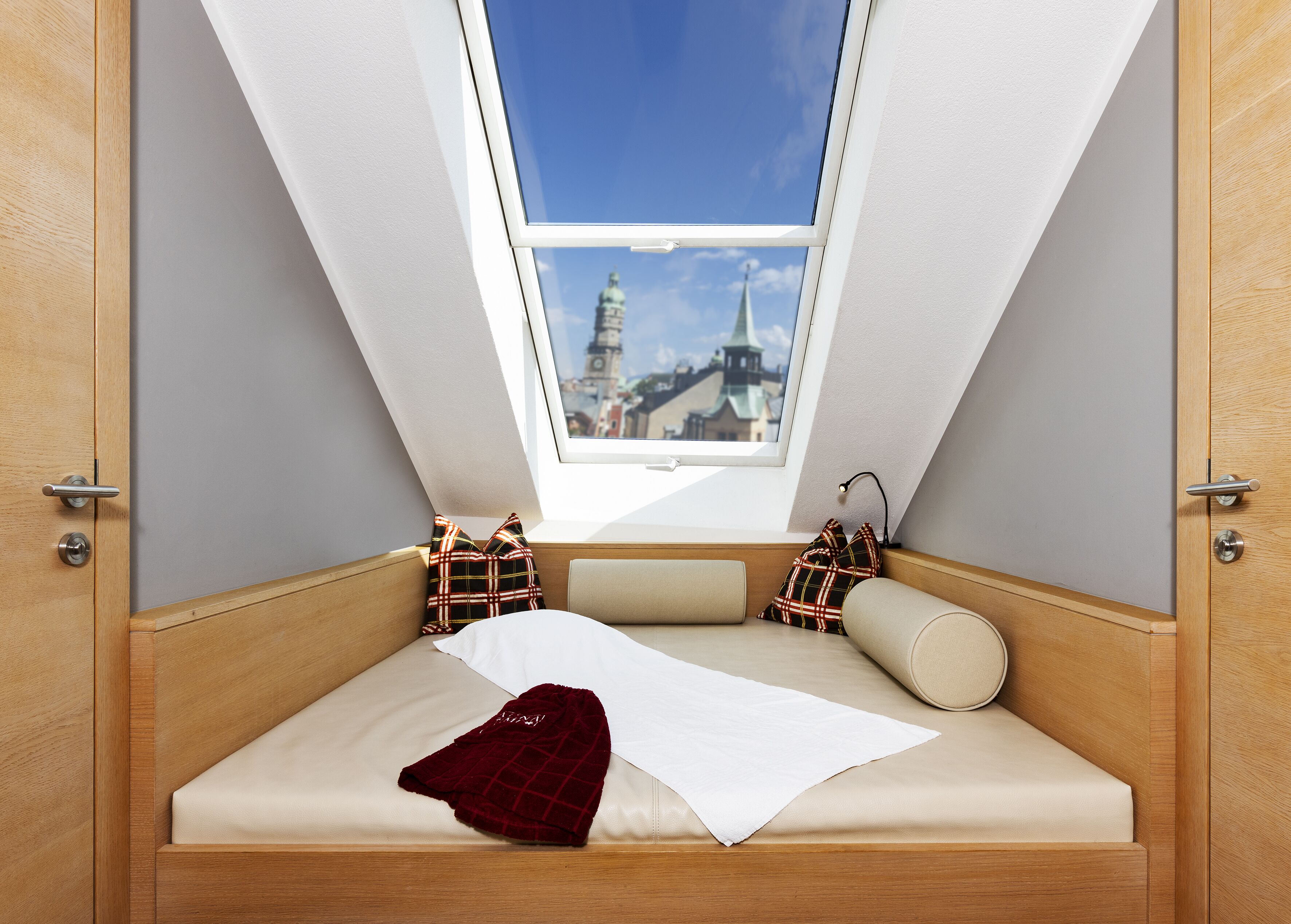 A room with a sloped ceiling, a bed with a white sheet, a red blanket, and plaid pillows.