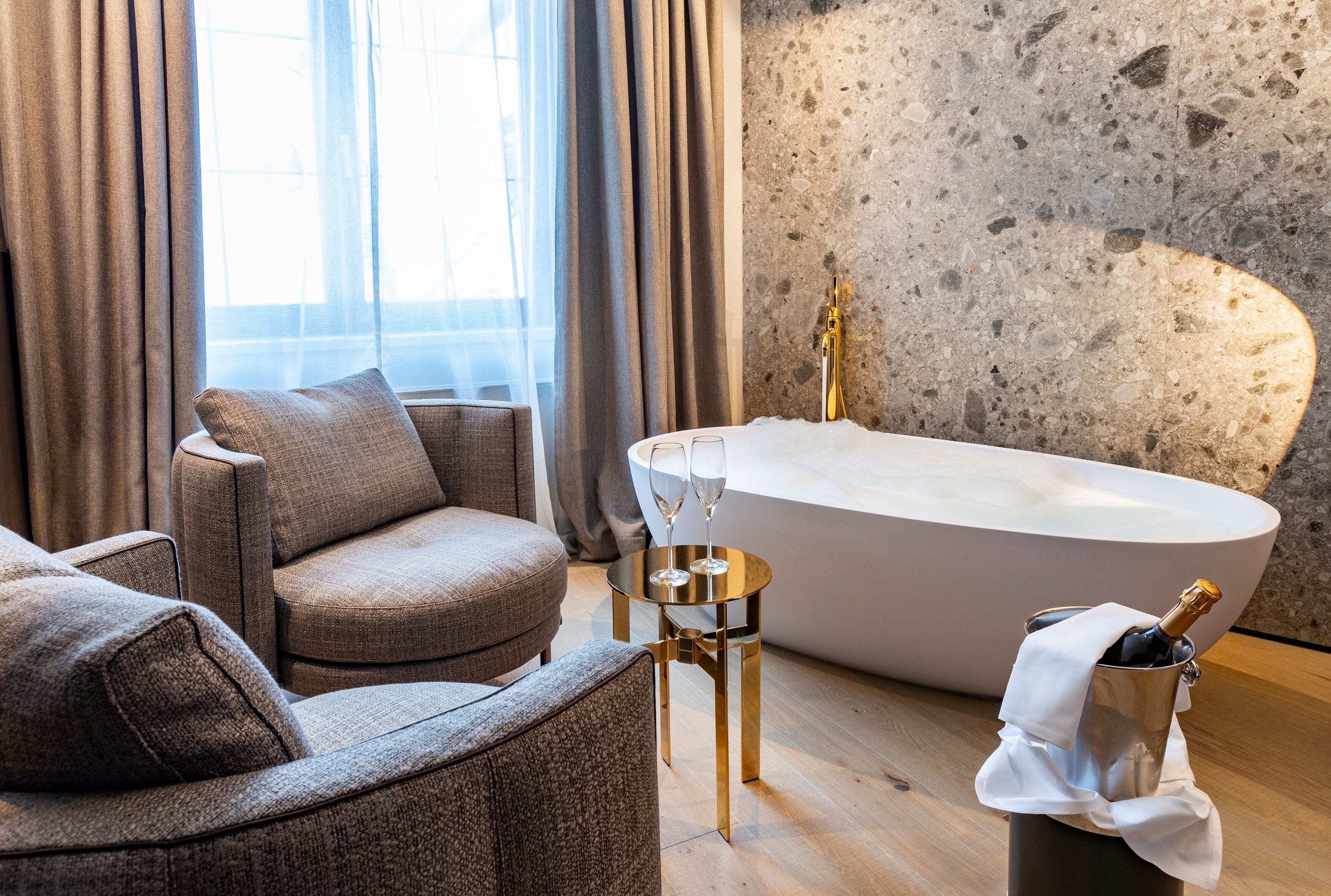 A modern bathroom with a freestanding bathtub, a round gold-framed side table, and a plush armchair.