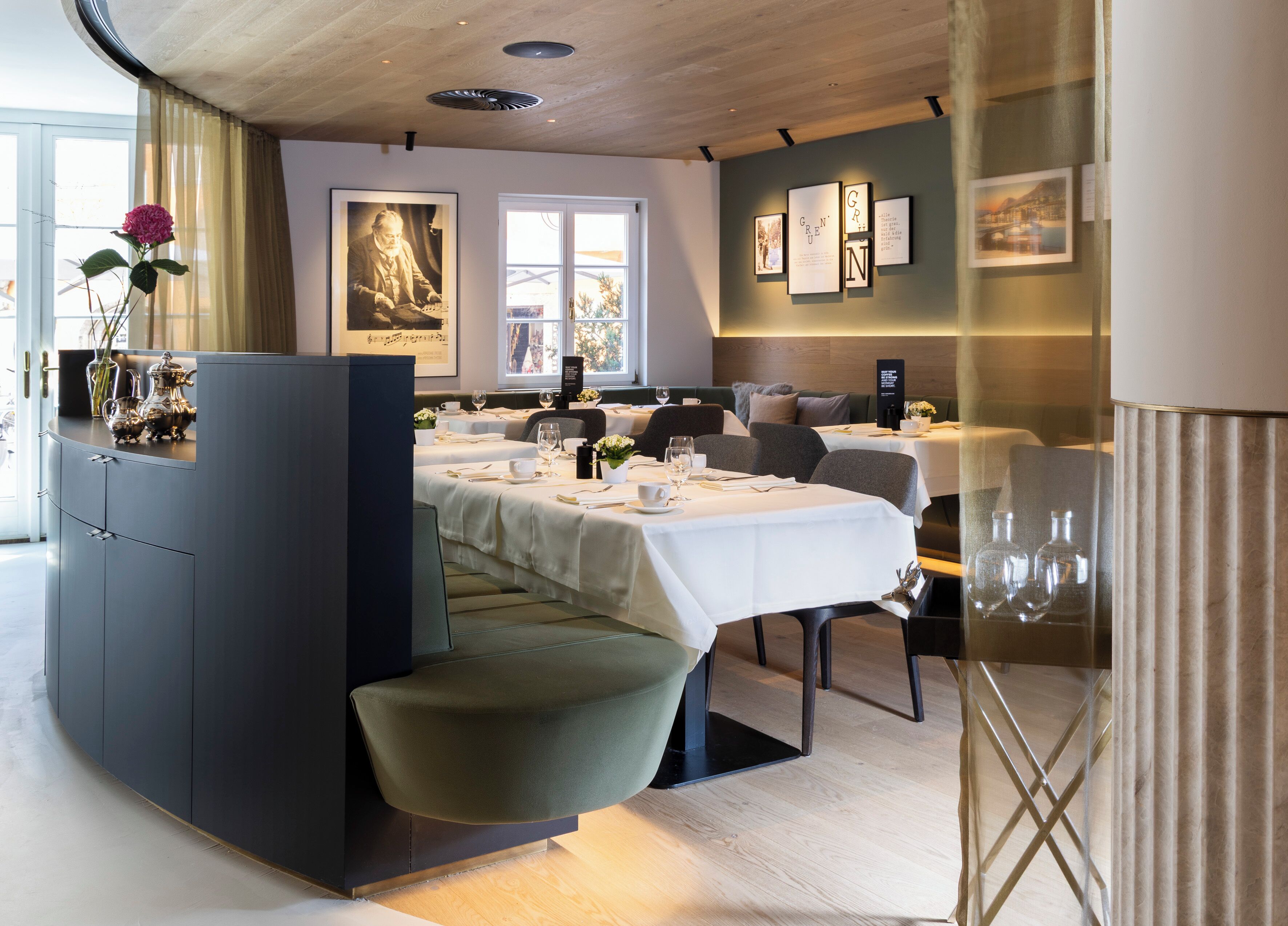 A restaurant with a modern interior, featuring a central dining area with white tablecloths, a dark-colored booth, and a vase with flowers on the left.