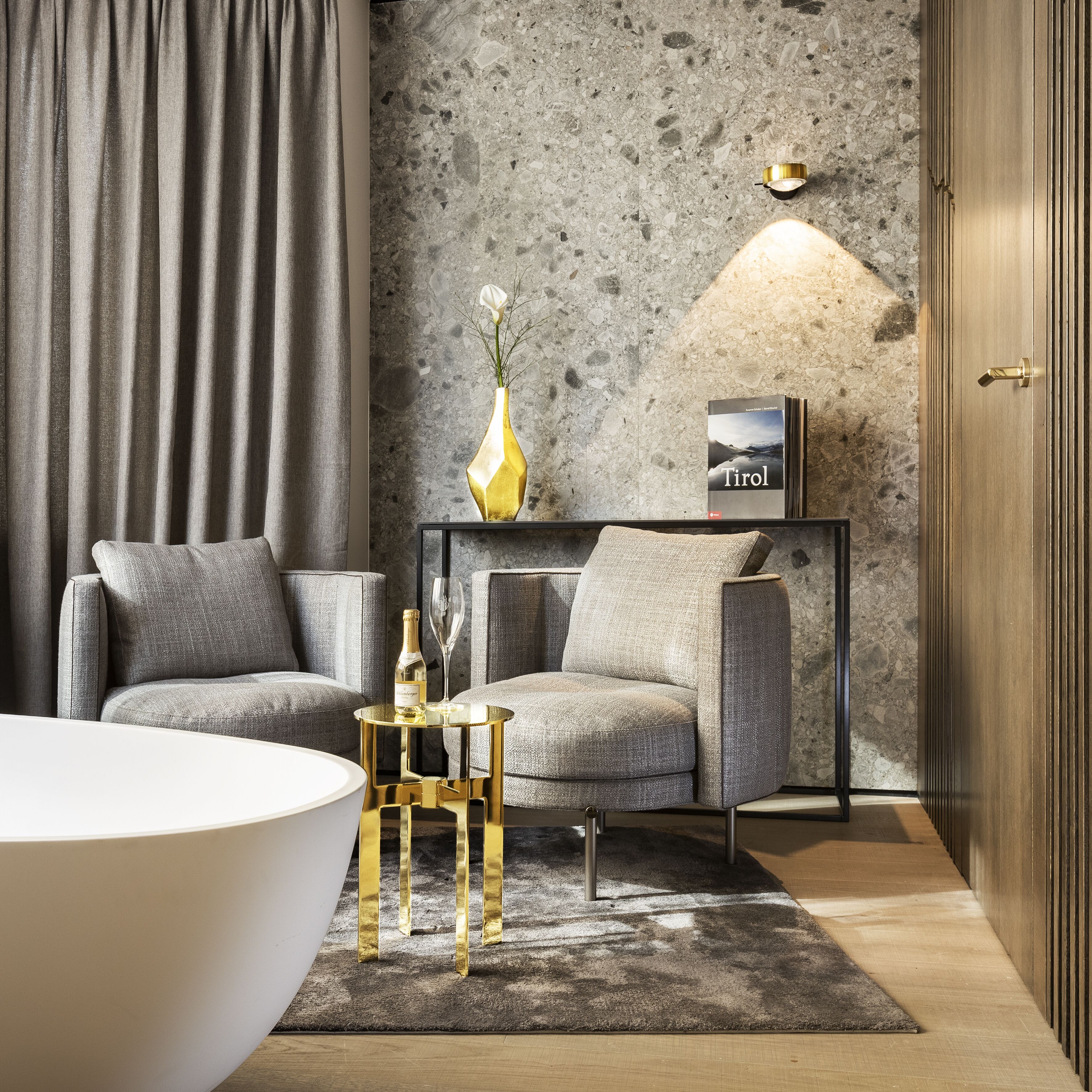 A modern living room with a grey sofa, a white coffee table, and a gold side table with a bottle and glass. A textured wall and a door with vertical lines are visible.
