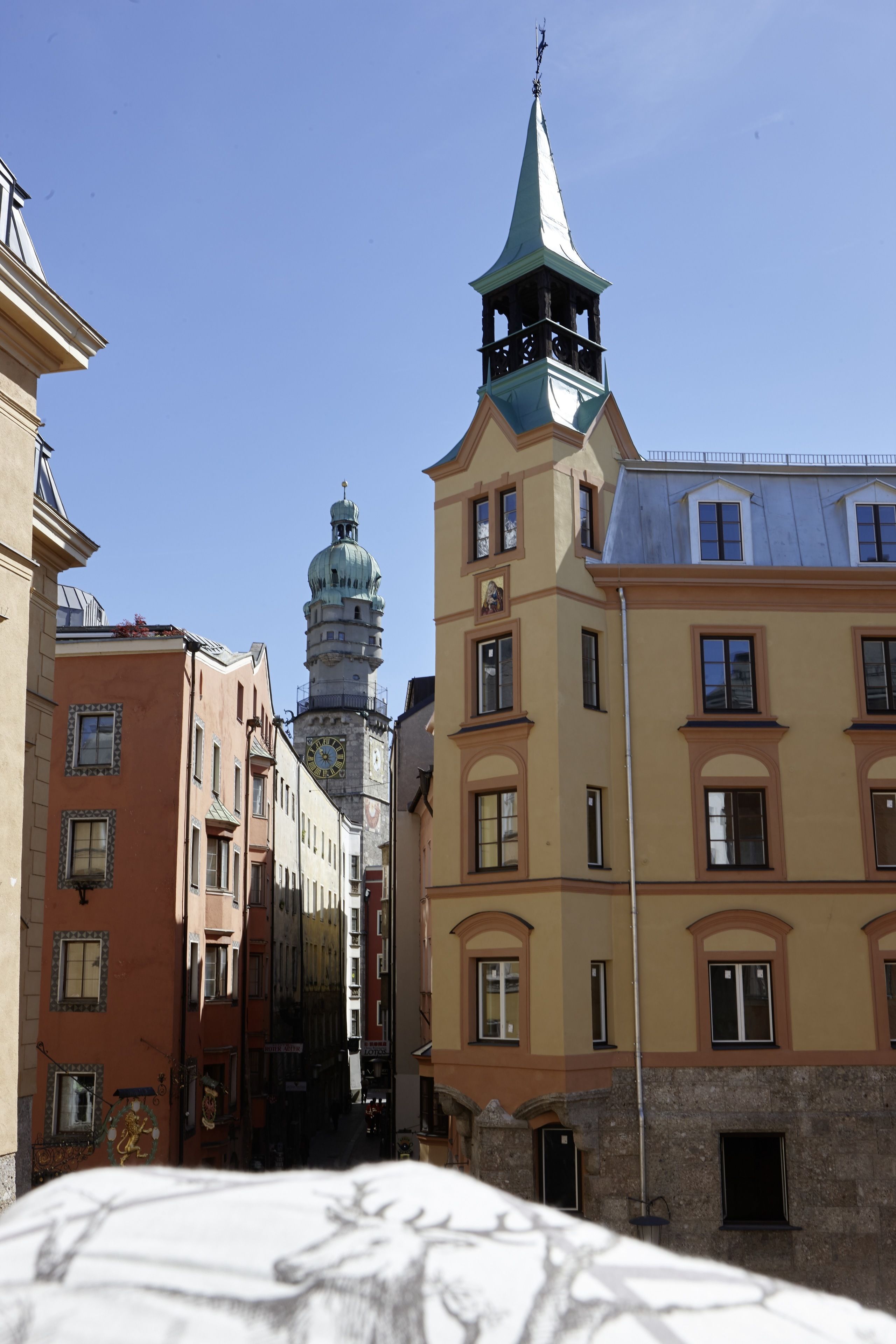 A narrow street with buildings on either side, a clock tower, and a church spire in the background.