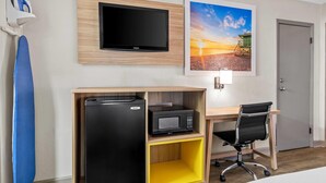Habitación de hotel con televisor de pantalla plana en la pared, refrigerador, microondas, escritorio con silla e imagen enmarco de una playa.