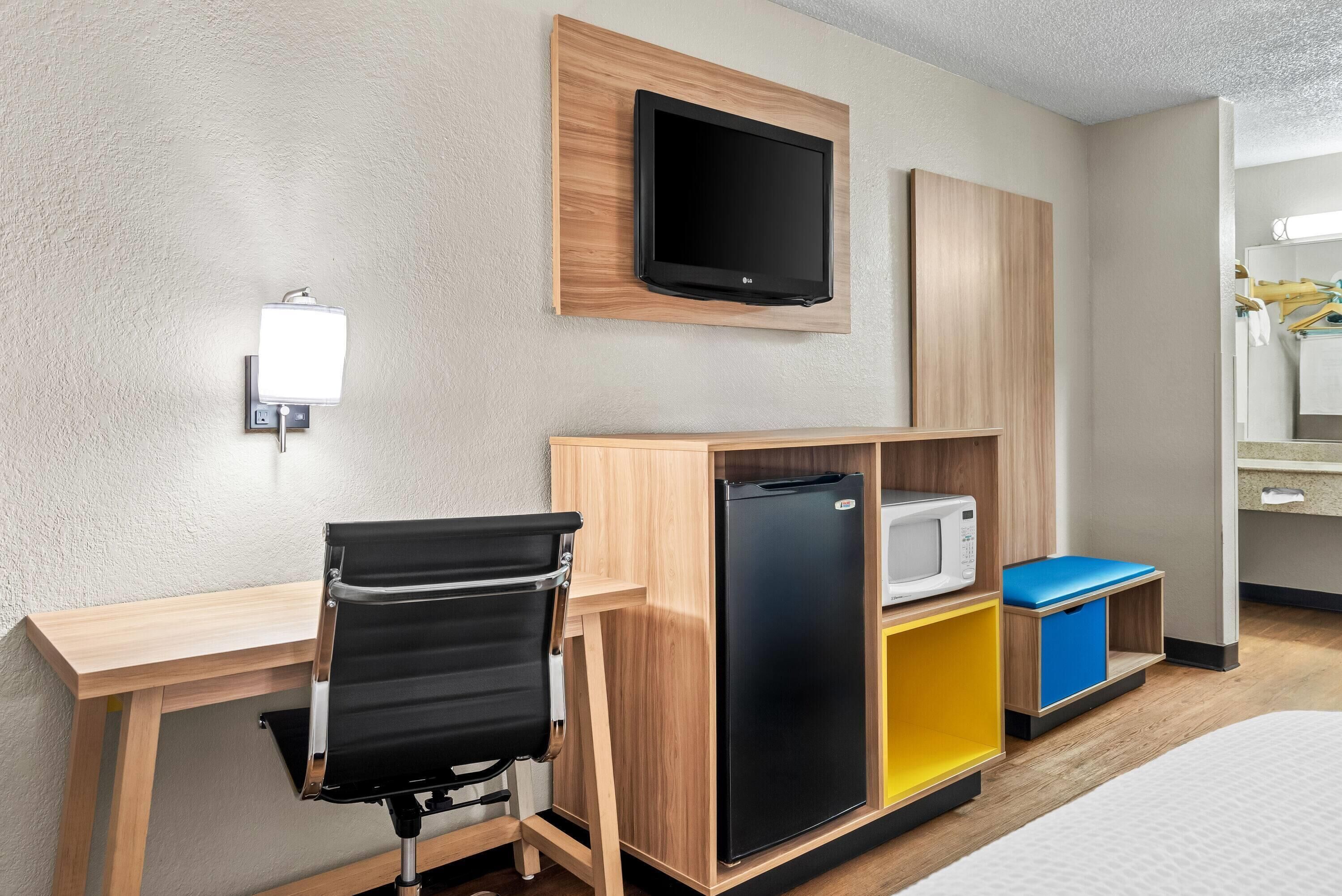 A hotel room with a wooden desk, a black office chair, a black refrigerator, a microwave, and a small blue storage unit.