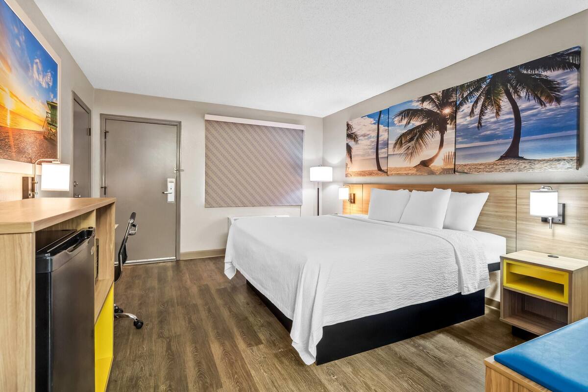 Ein Hotelzimmer mit Bett, Nachttischen, einem Schreibtisch mit Lampe, einem wandmontierten Fernseher und einem großen Gemälde im Strandhausstil.