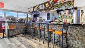 Zona de bar con pared de piedra, barra de madera y varios taburetes. Se observan varias botellas y vasos sobre la barra, así como una grifería de cerveza grande.