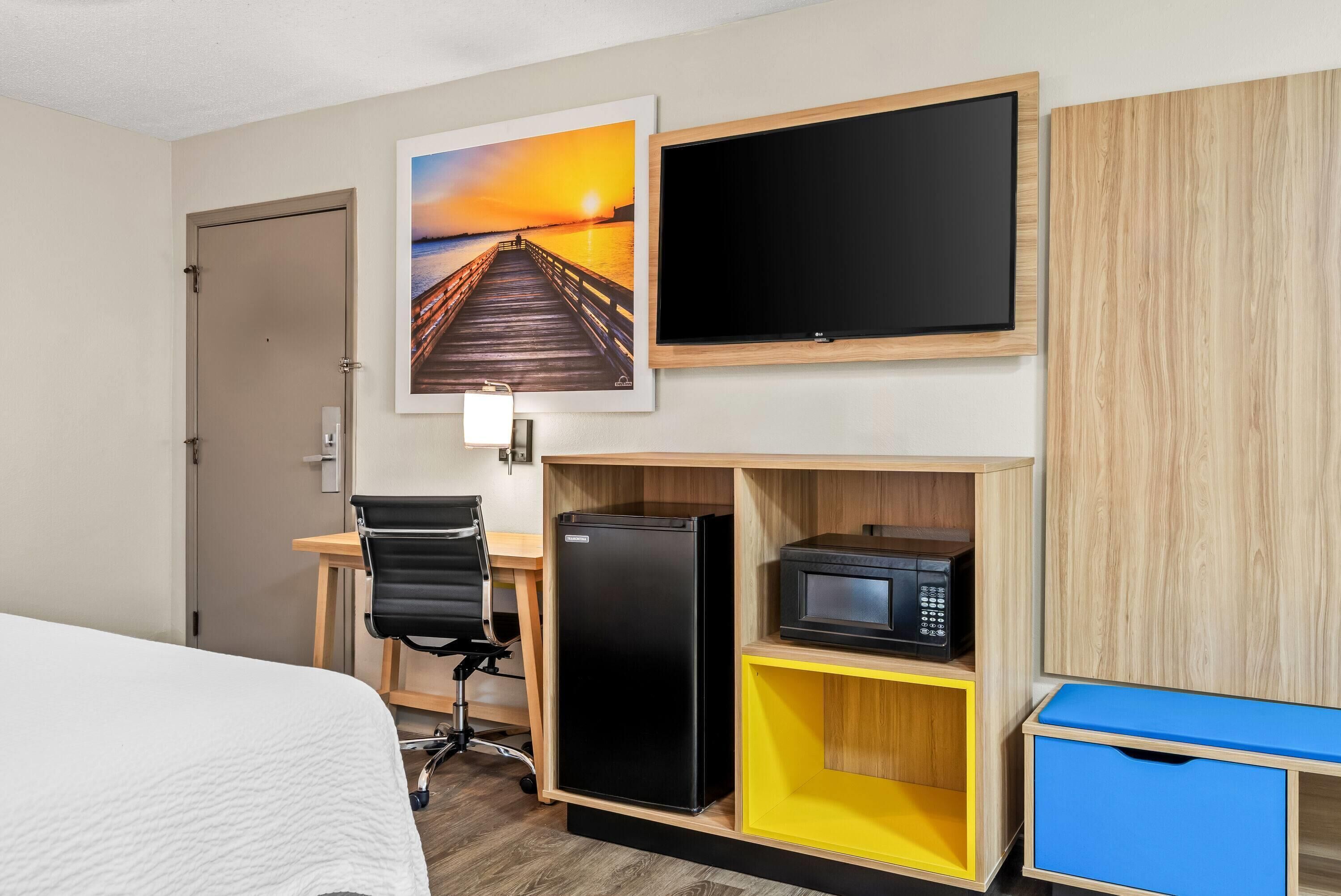 A hotel room with a bed, a desk with a chair, a black refrigerator, a microwave, a TV, and a wooden cabinet.