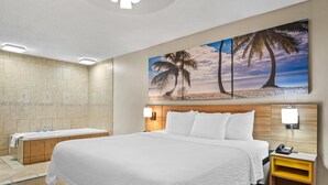 Habitación de hotel con una cama grande, cabecera de madera, un cuadro de palmeras, una bañera y una mesita de noche con lámpara.